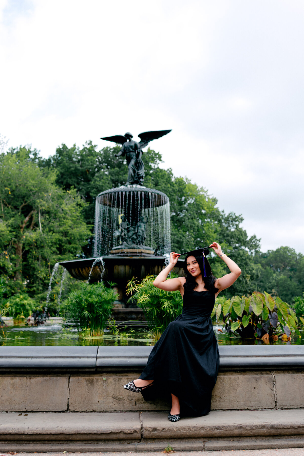 NYU graduate wearing black dress taking playful photos with cap at Central Park.