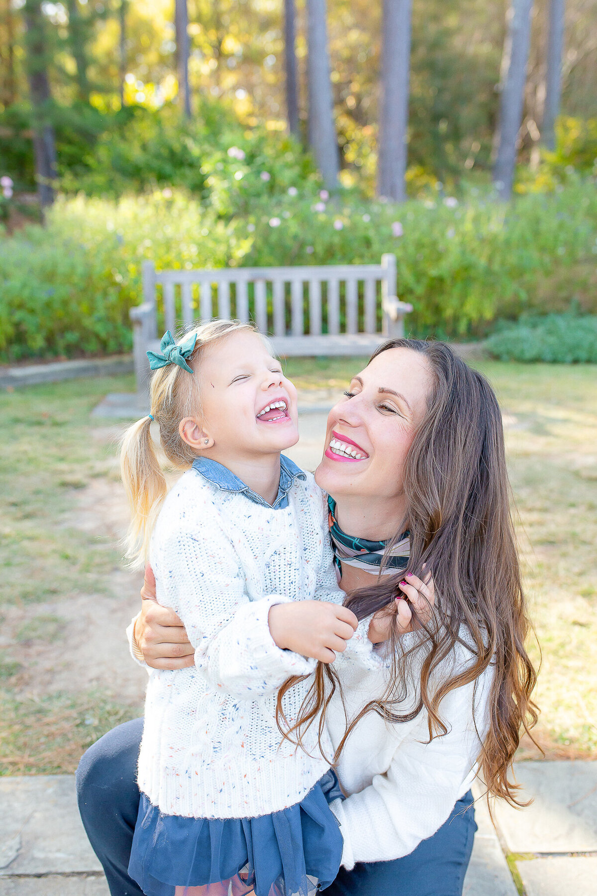 A mother and her young daughter share a joyful moment outdoors, laughing together in the golden sunlight. Dressed in cozy sweaters with a garden backdrop, this heartfelt portrait captures the warmth and love of family in a Birmingham family photography session.