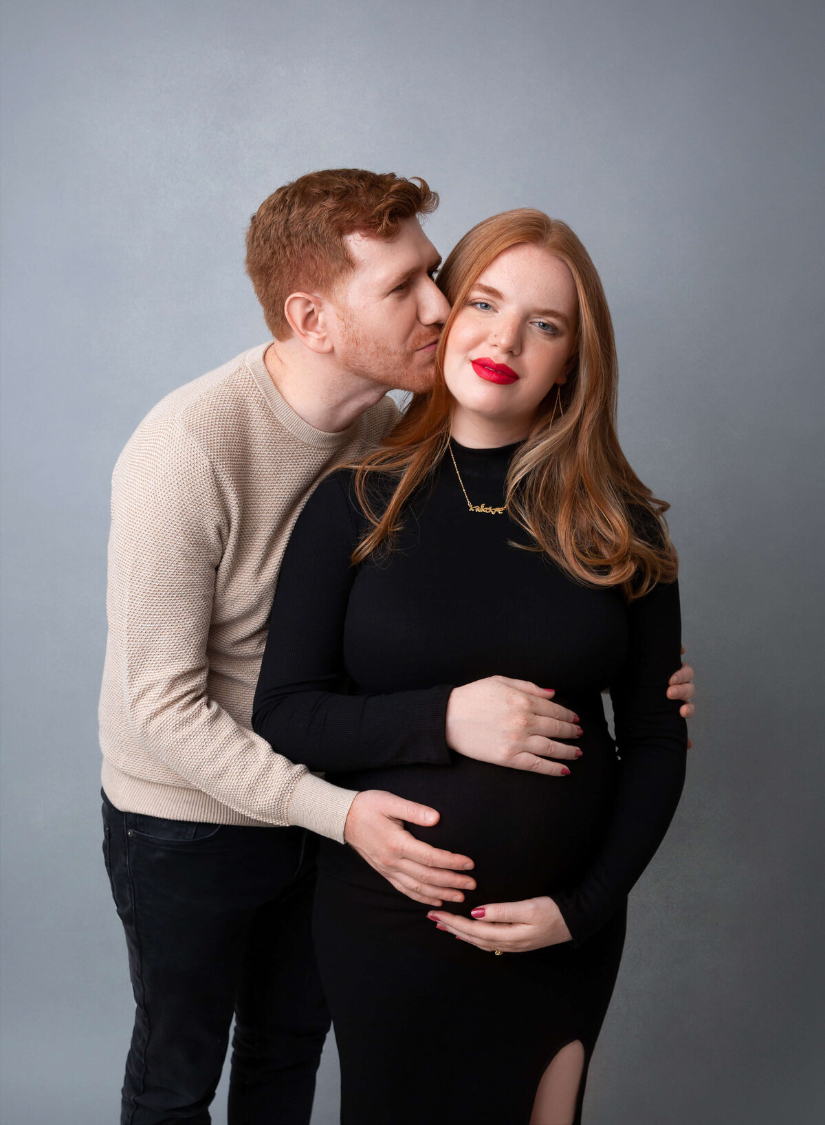 A man leans over kissing his pregnant wife in a black maternity one piece maternity suit after maternity pictures in NYC