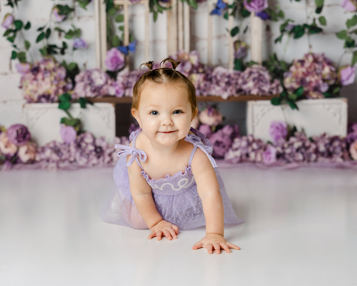 Baby crawling in lavender dress with purple flower backdrop during cake smash session. Photograph by Yucaipa cake smash photographer Kaitlyn Dawn Photography.