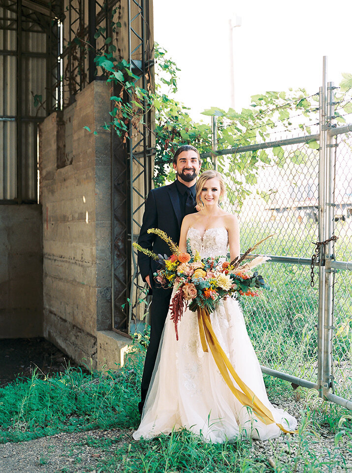 Bride and groom, wearing a black tuxedo and white wedding gown, smile with a large bouquet of yellow, blush and orange flowers