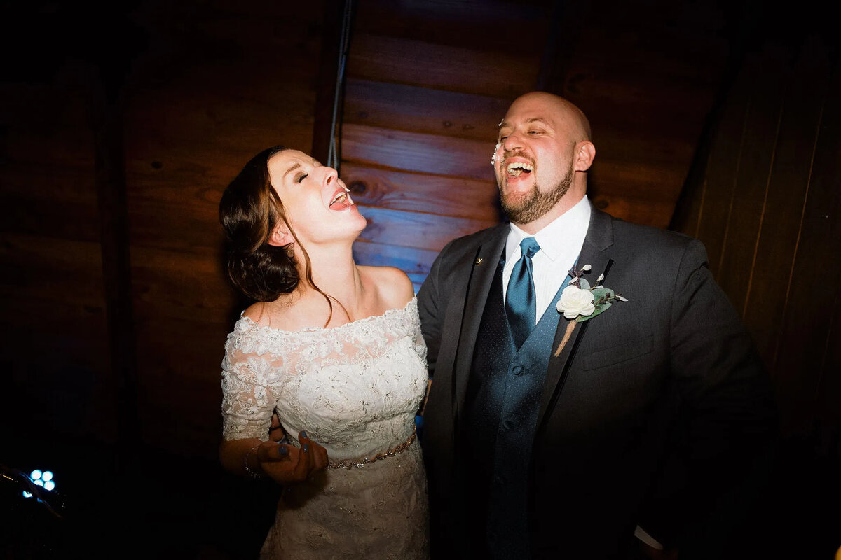 A bride and groom in formal attire laugh joyfully together under warm lighting, standing close in front of a wooden wall at their wedding celebration, beautifully captured by an NJ wedding photographer.