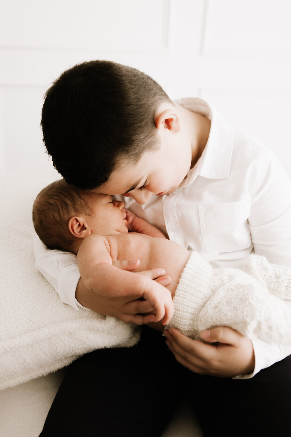 Older brother gently cradling his newborn sibling, their foreheads touching in a quiet, tender moment during a newborn session in a softly lit Sydney studio.