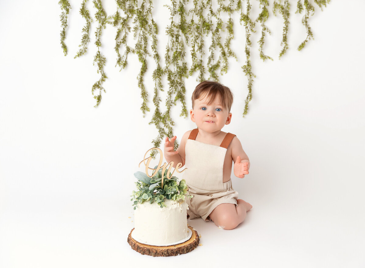 Baby in canvas overalls sits in front of a cake for is first birthday cake smash photoshoot. White cake is topped with greenery. Baby is looking at the camera.