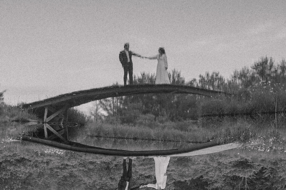 A bride and groom stand hand in hand after their wedding ceremony at  Blenheim Hill Farm on a wooden bridge, their reflections perfectly mirrored in the calm water below. The black and white photo adds a timeless, nostalgic feel to this romantic and serene moment, emphasizing the couple's connection and the beauty of their surroundings.