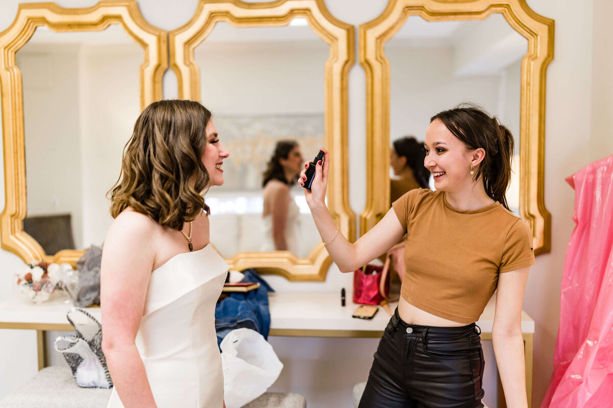 A smiling makeup artist wearing a brown crop top and black pants applies makeup to a woman in a white strapless dress in front of two gold-framed mirrors.