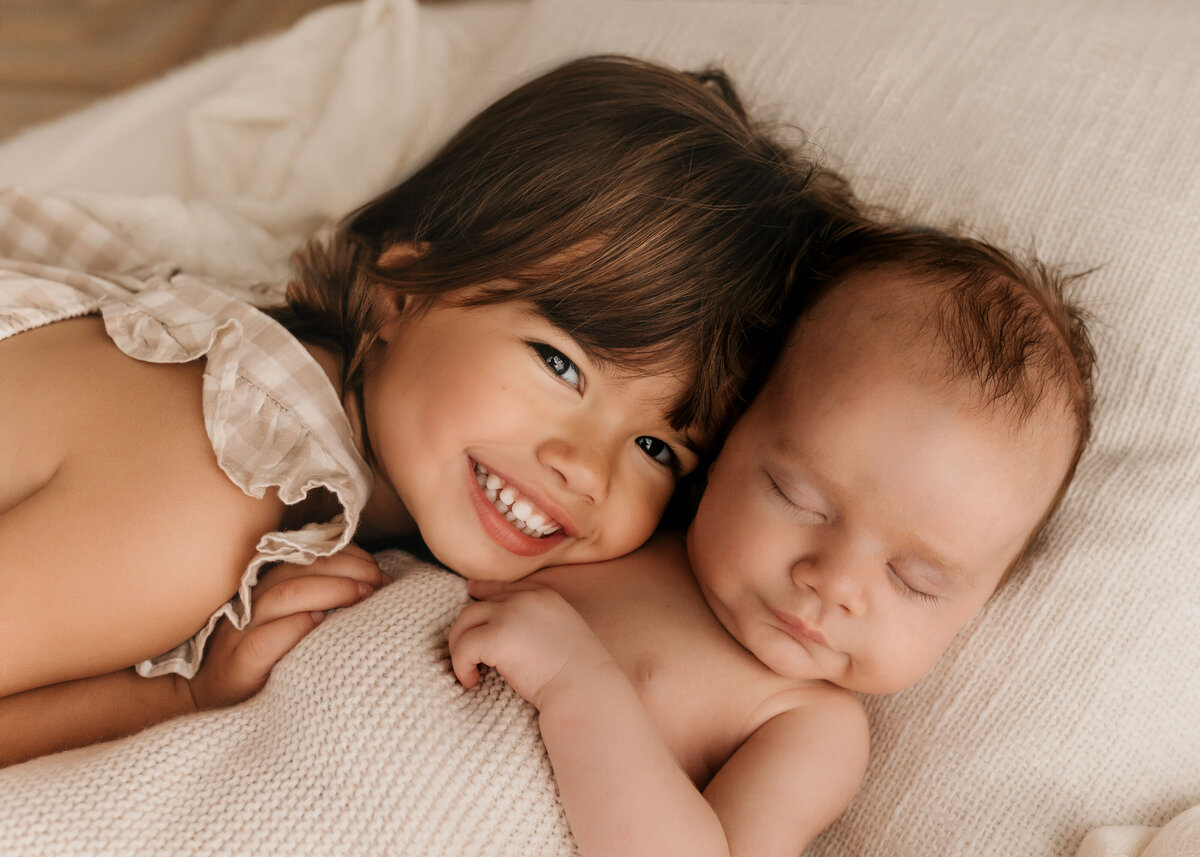 Photo of a newborn baby sleeping whilst his older sister cuddles him and smiles at the camera during a photoshoot
