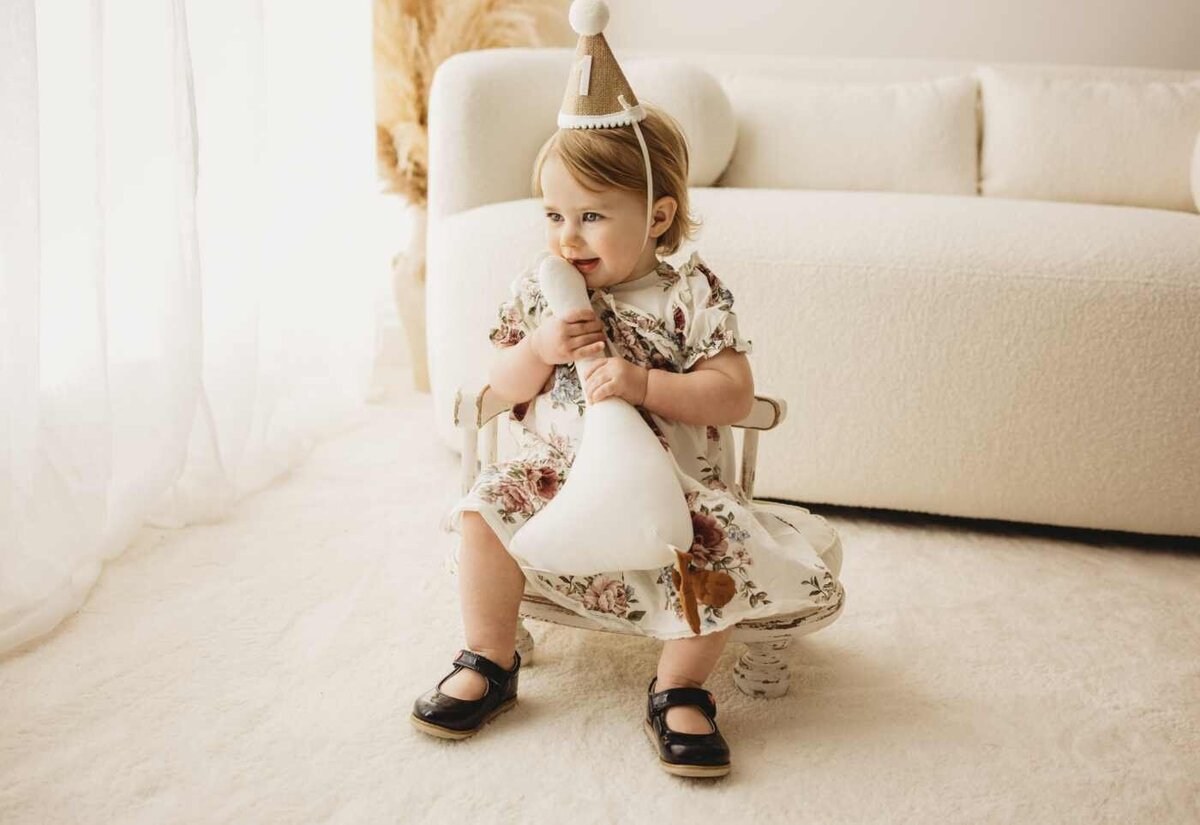 Ruua Hamdan Photography (UK): A baby in a floral dress and party hat sits and holds a white toy