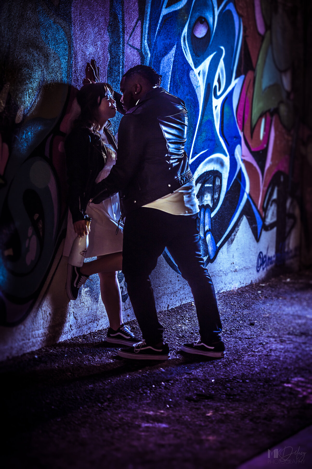 las vegas graffiti alley  groom leans in for kiss with bride pressed back against the wall  bride in white dress with black leather jacket grom in skinny jeans and white dress shirt with black leather jacket wearing  converse on Fremont st  las vegas elopement eloping in vegas  las vegas wedding photographers las vegas wedding photography mk delacy photography