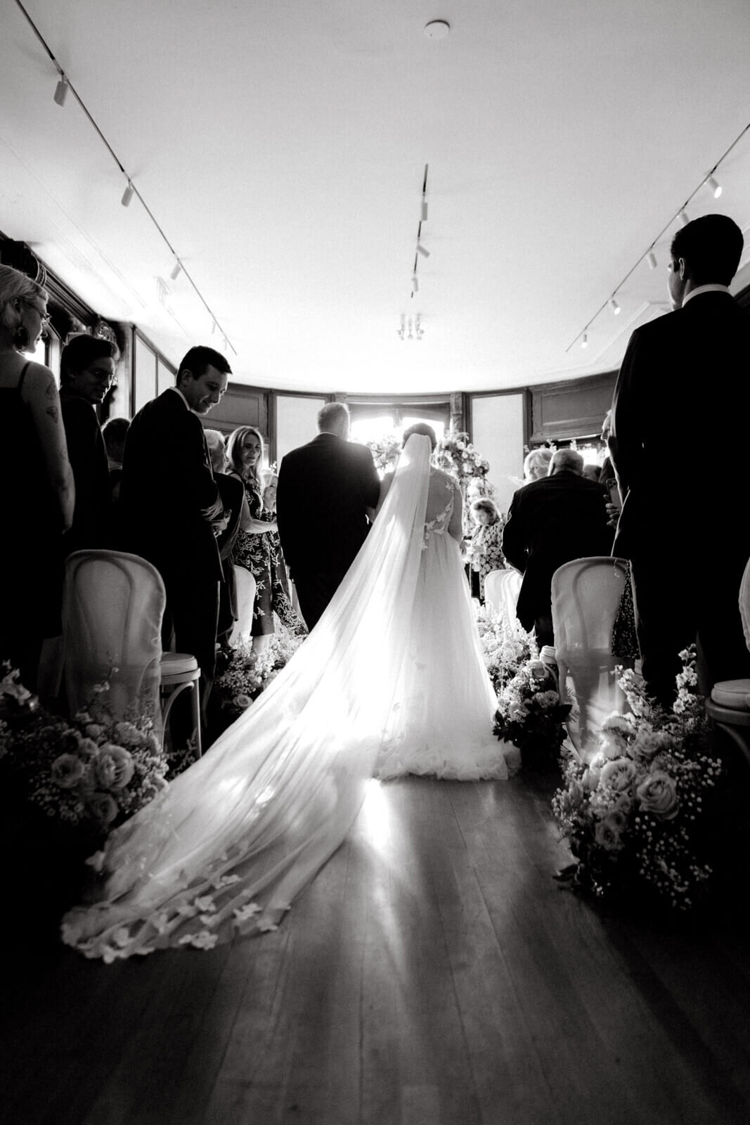 Black and white photo of the bride and the groom, standing on their backs, with people standing on both sides.