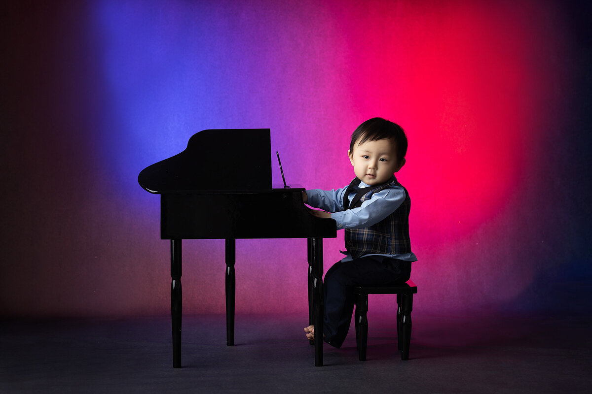 Dallas milestone photographer captures baby playing piano in formal outfit with colorful backdrop.
