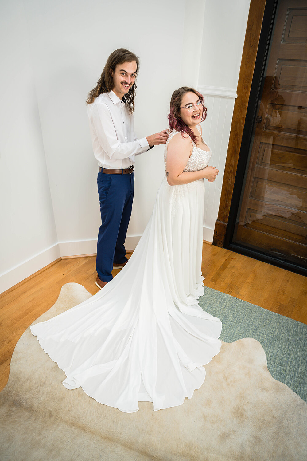 A marrier helps his partner get into her wedding dress by buttoning the back of her gown in a hotel room at Fire Station One in Roanoke, Virginia. The two smile upwards at the photographer (not photographed) taking their photo.