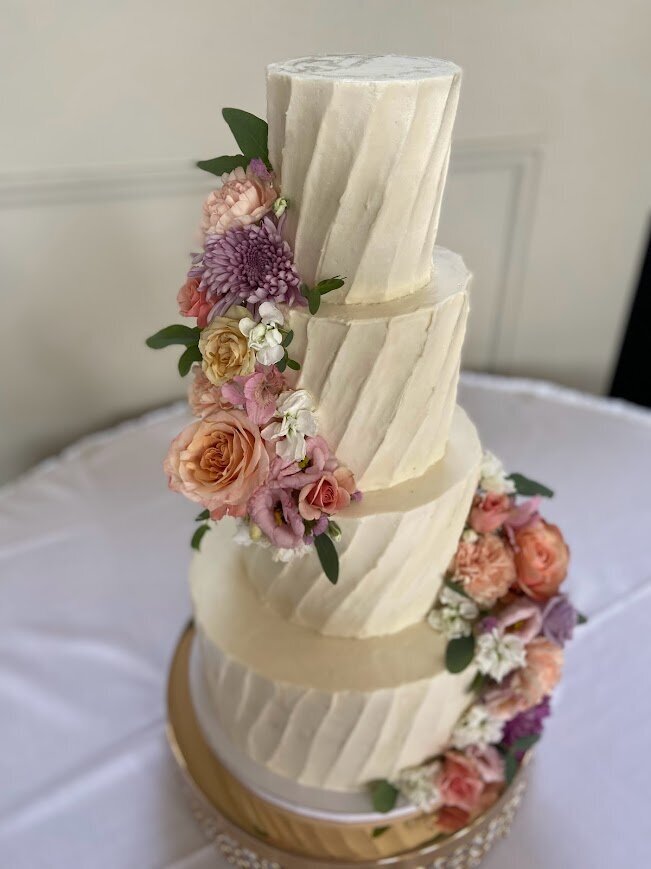 Photo shows a four tier cake with white frosting in a swirl lined pattern. On the bottom right and on the top left are large clusters of fresh flowers in orange, pink, and purple. 