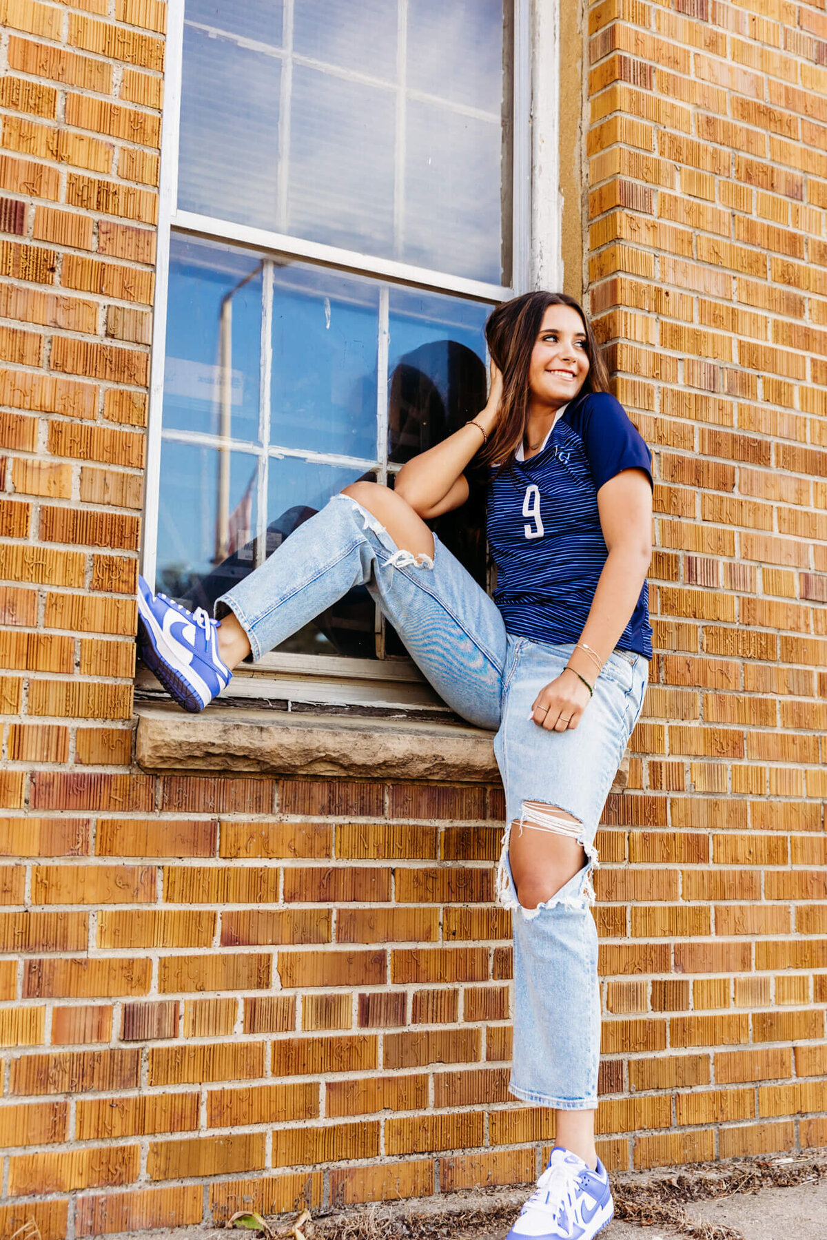 young woman laughing as she sits in a window sill while resting her elbow on her knee and looks out into the street 