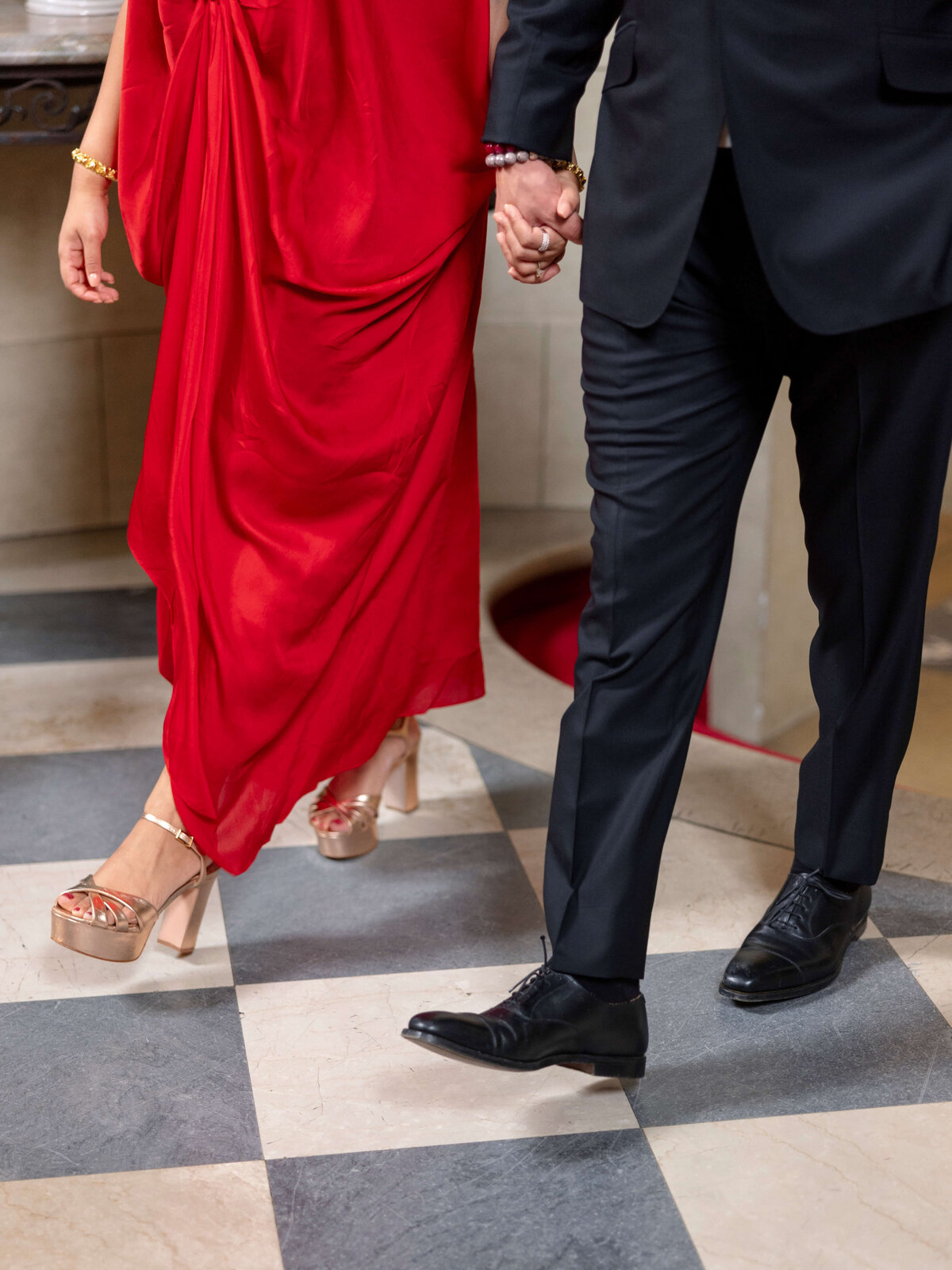 A couple holding hands walks on a checkered floor. The woman wears a long red dress and platform heels, while the man is in a dark suit and black shoes.