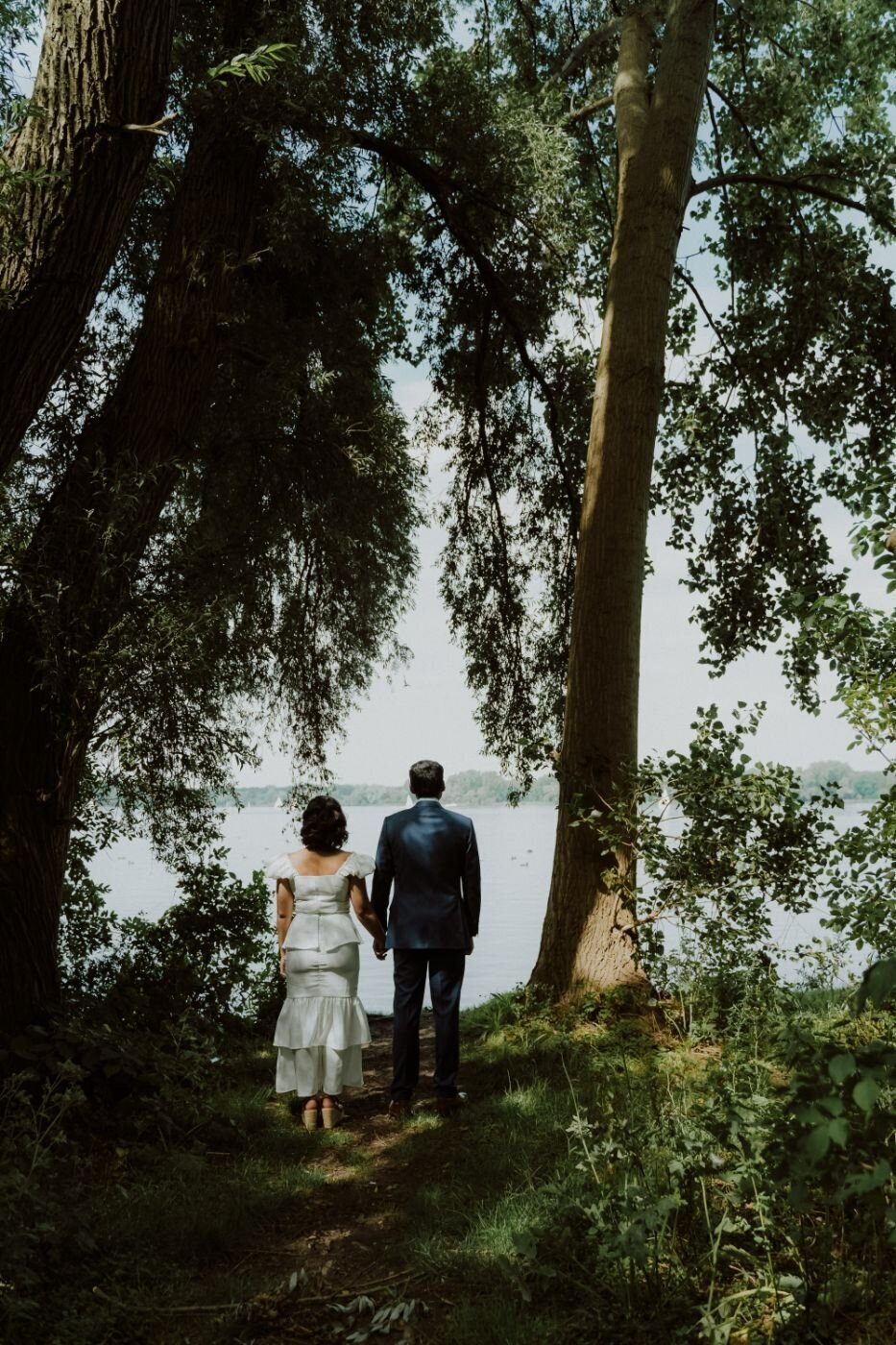 A romantic shot of a couple walking hand-in-hand down a long, white wooden bridge that extends over a lake. They are smiling and looking at each other. The man is wearing a white shirt and blue pants, and the woman is wearing a colorful patterned dress. Trees and lush greenery line the edges of the water.