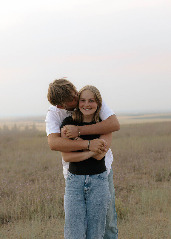 During a couples photography session in Rexburg, ID, a man holds his girlfriend from behind. He's kissing her on her temple as she smiles at the camera.