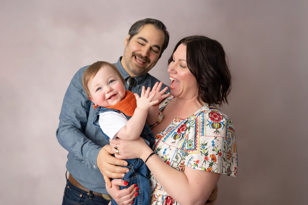 Parents smile in awe looking at their baby who is clapping while mom is carrying him.