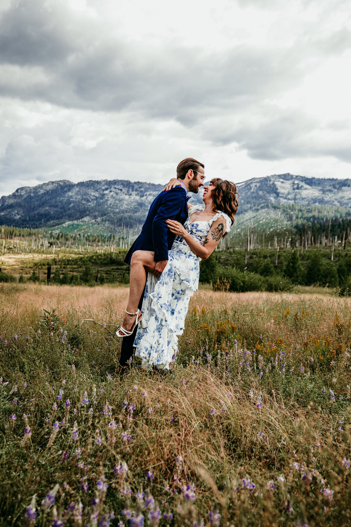 Bride and groom sharing a playful, romantic moment in a wildflower meadow near Lake Tahoe, with dramatic mountain views under a cloudy sky.