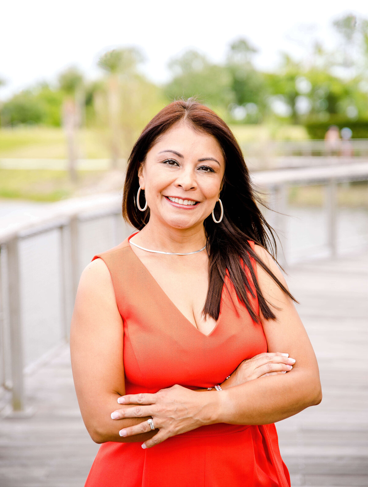 Brand photographer near me photographs woman for an outside ran session standing on a dock on a lake with her arms crossed while smiling wearing a red blouse and gold hoop earrings for her brand photo headshot