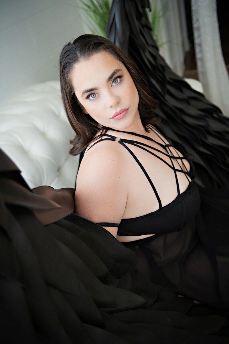 A woman with striking blue eyes and dark hair leans on a white couch. She wears a black strappy outfit and is wrapped in flowing black fabric.