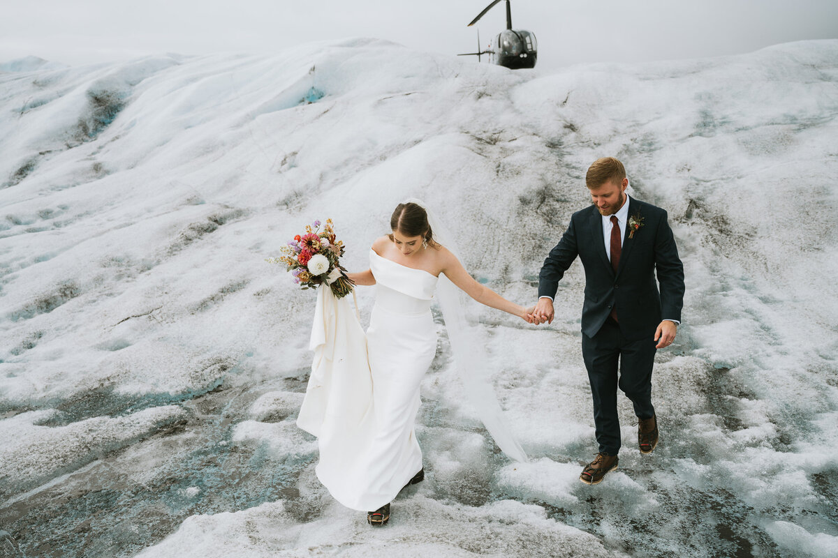 couple in wedding attire walking across glacier ice from the helicopter holding hands and a colorful bridal bouquet