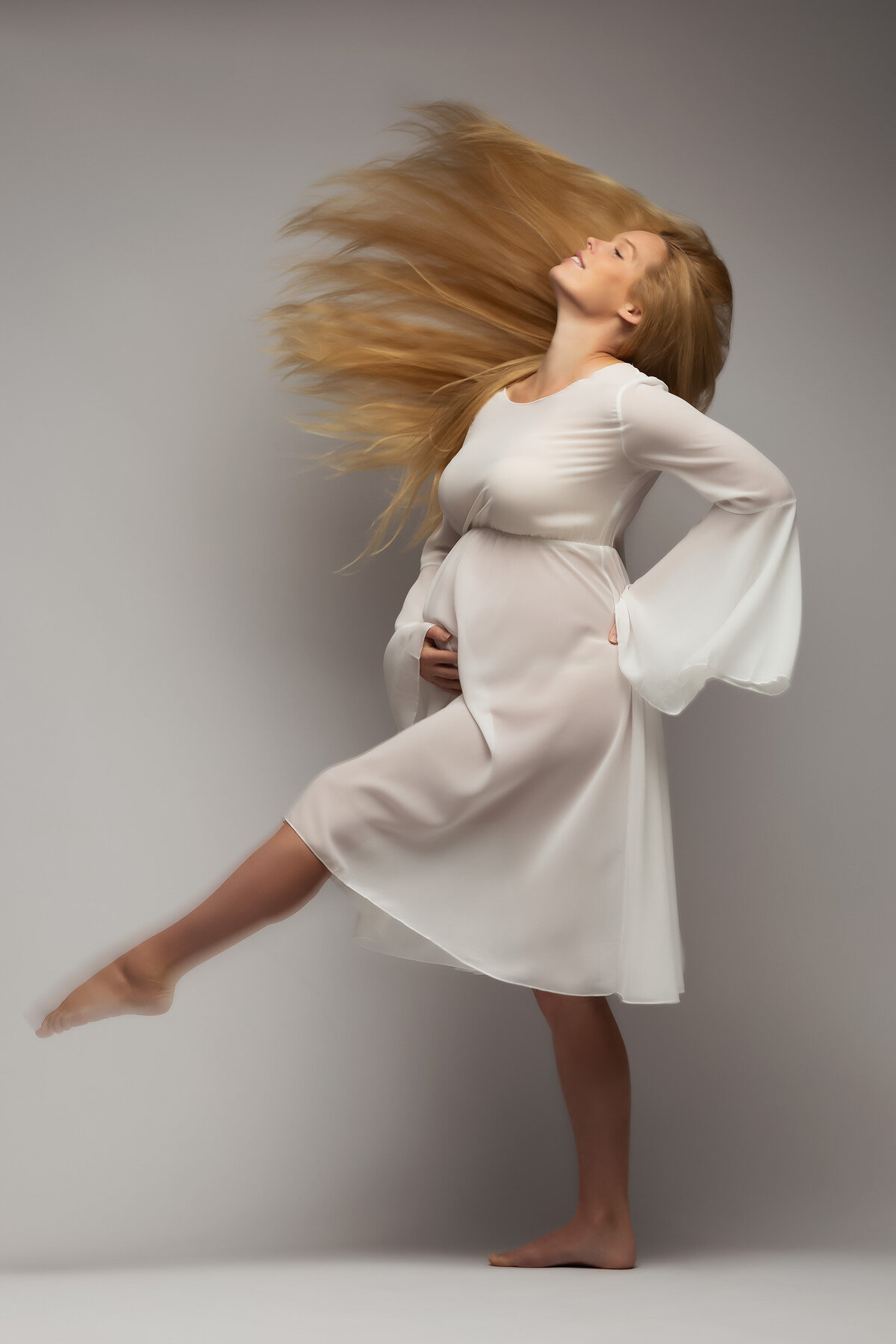 A woman gracefully tosses her long blonde hair while wearing a flowing white dress, exuding pure joy and elegance. Her carefree pose captures the artistry of maternity photography, perfect for celebrating the beauty of DFW maternity photography.