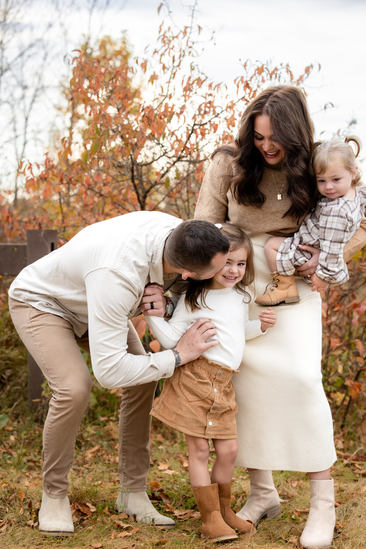 a father leans down to kiss her daughter as she laughs, while a mother holds onto their other child. the family is wearing neutral fall clothing