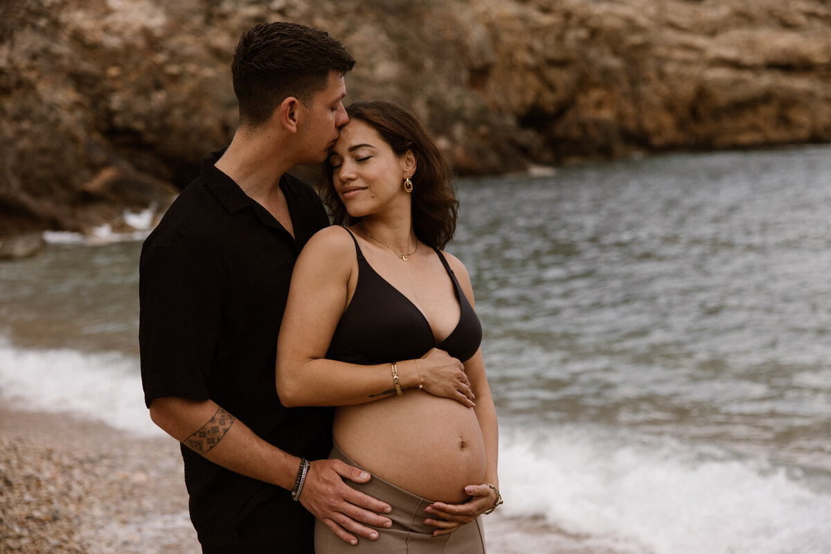 A man is standing behind a woman and kissing her on the forehead on a beach on Ibiza for a pregnancy photoshoot