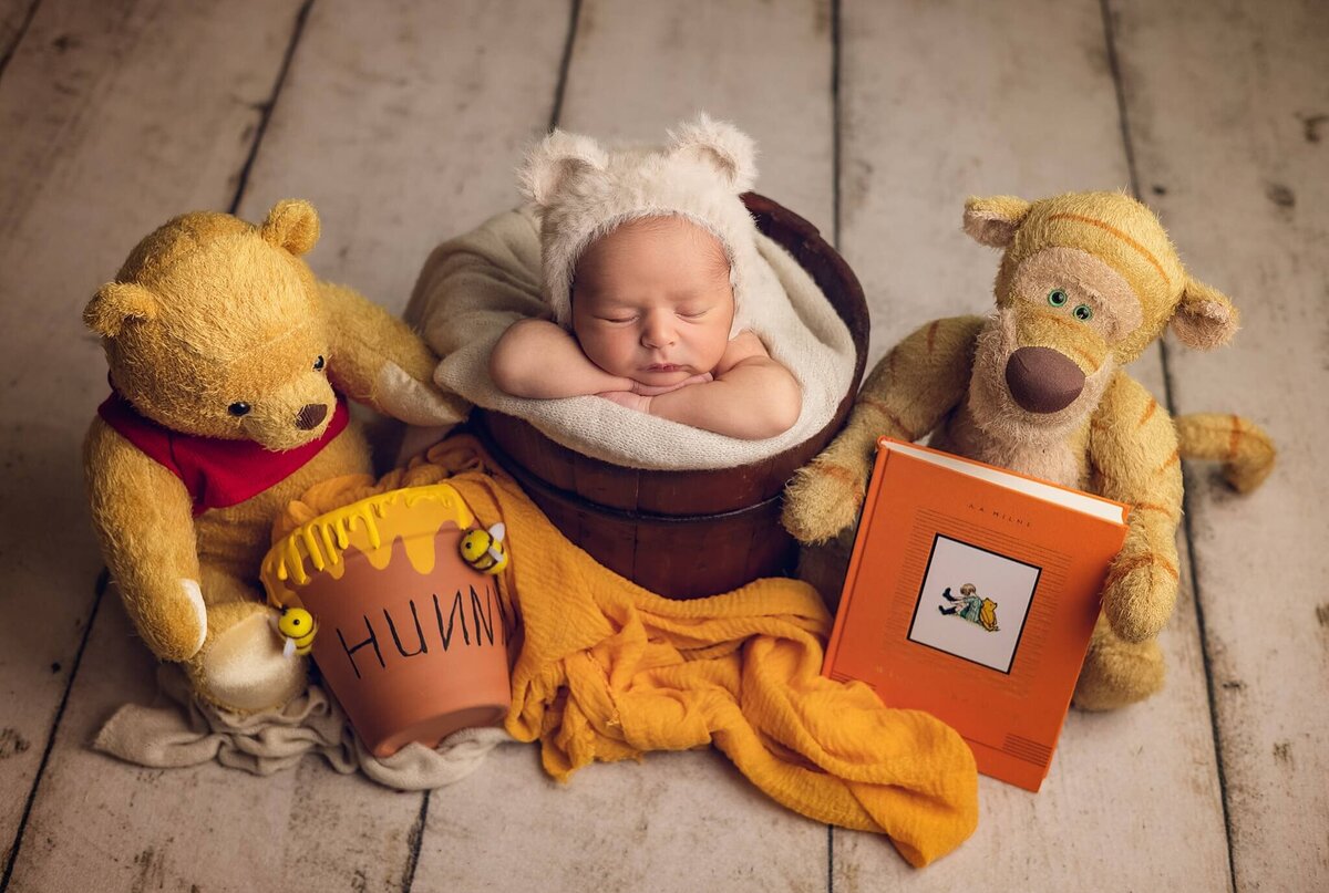 Baby sleeping in a wooden bucket surrounded by a plush Winnie the Pooh bear, a plush tiger, a "Hunny" pot, and a book, on a wooden floor.