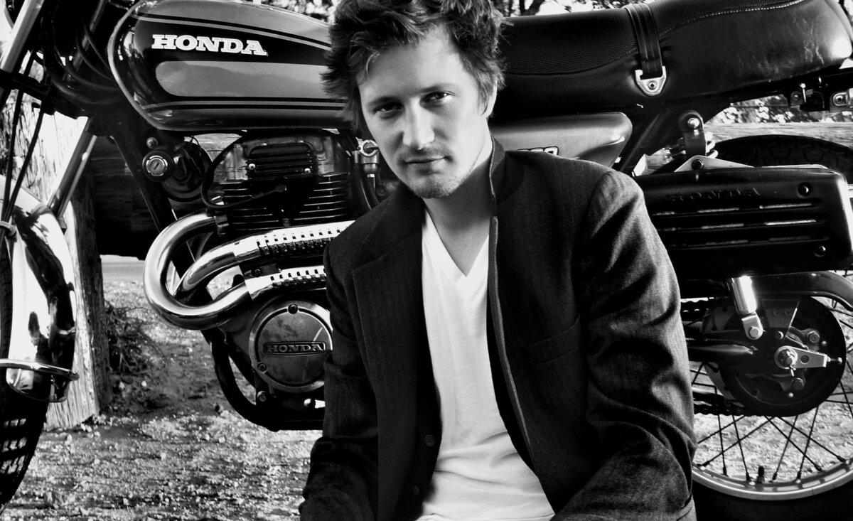 Male musician photo black and white Del Barber wearing white t shirt black suit jacket leaning against vintage motorcycle