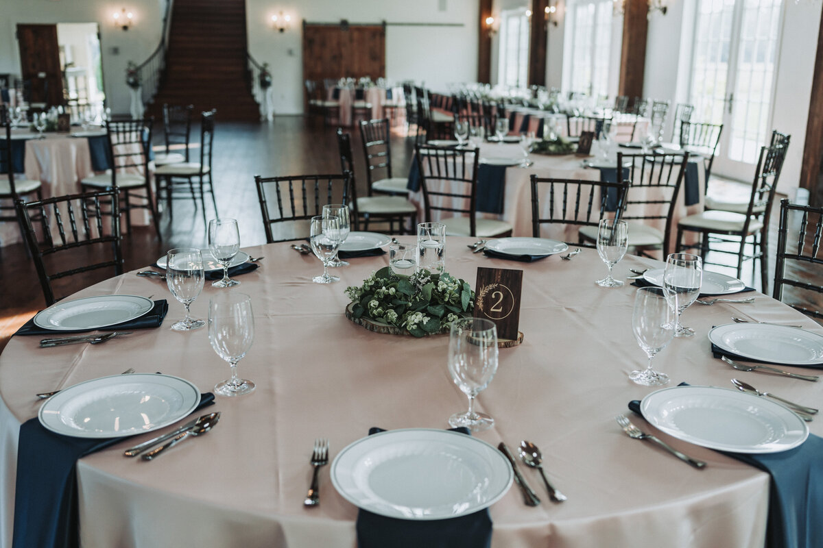 A beautifully arranged wedding reception table at Burlington Wedding and Events Venue, featuring a round centerpiece of greenery and white flowers, set with elegant plates, silverware, and glasses. The venue includes large windows, wooden floors, and a staircase in the background. Shot by Quinn Photography LLC.