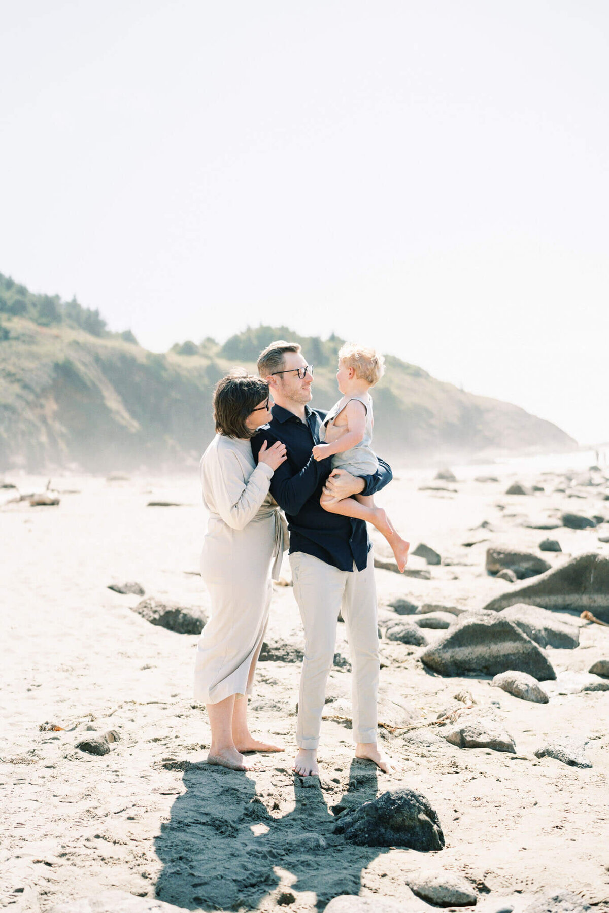 light and airy beach photos of a dad holding toddler with the forested cliffs of the oregon coatline in the background
