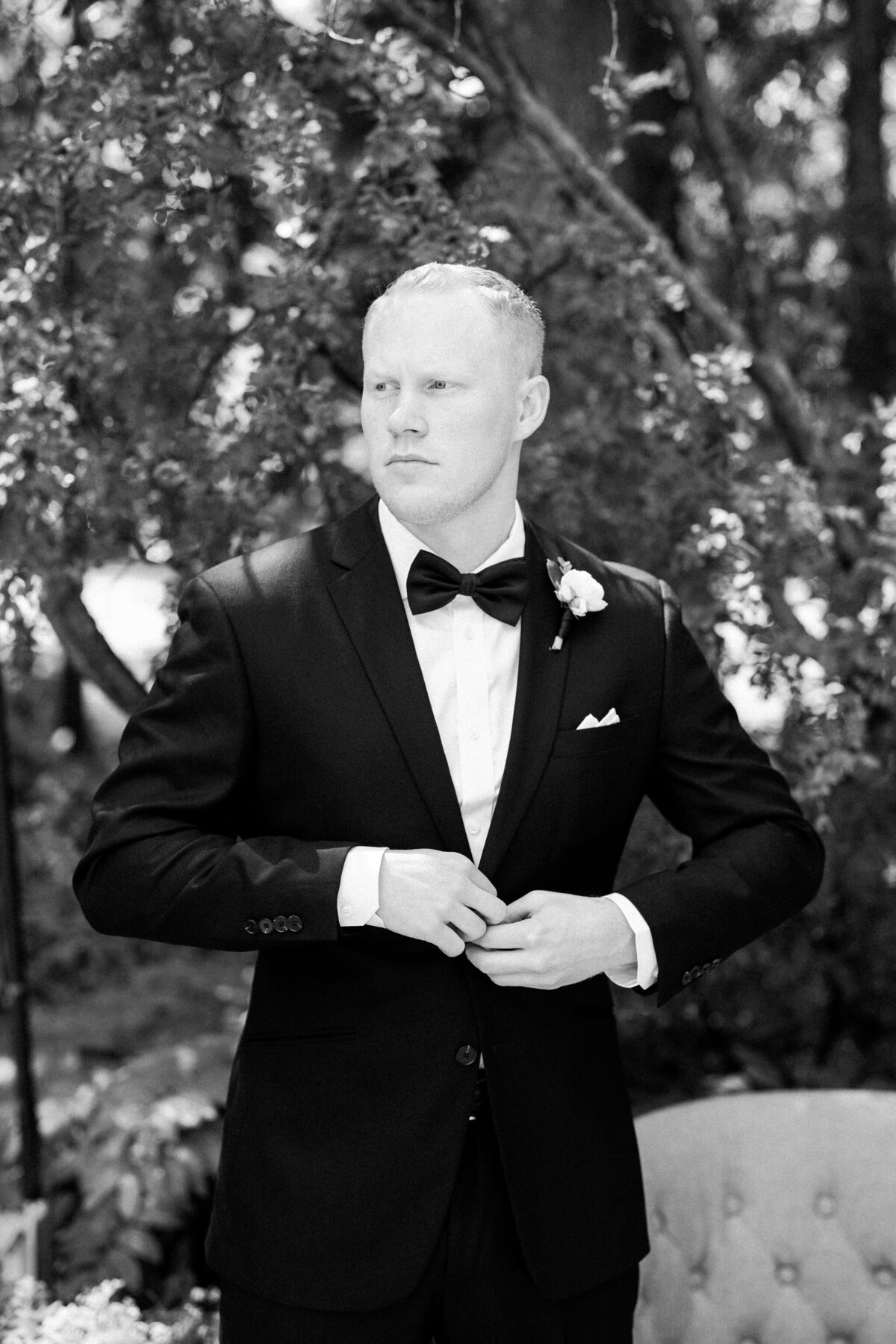 A man in a black suit and bow tie stands outdoors adjusting his suit jacket, looking off to the side with trees in the background, capturing a serene moment at a wedding in Calgary.