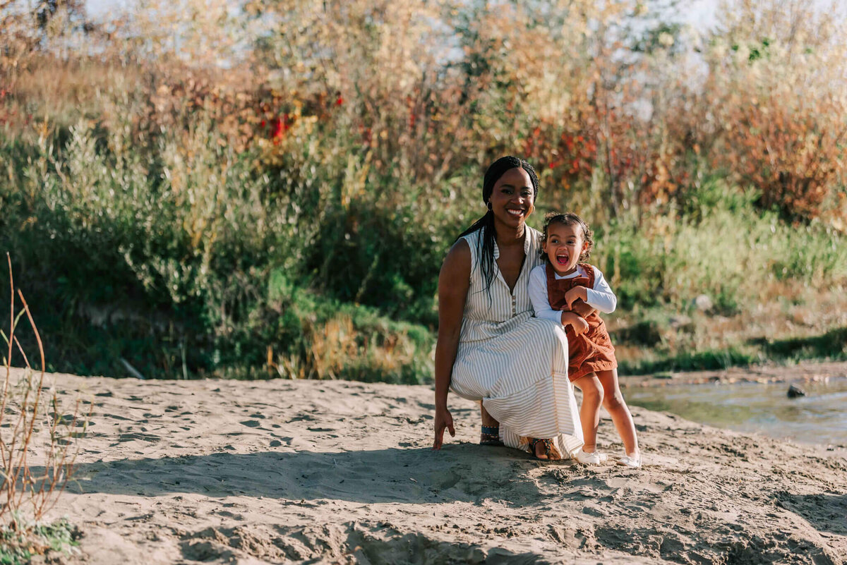 Mom is kneeling on the sand and the little girl stand by her. 