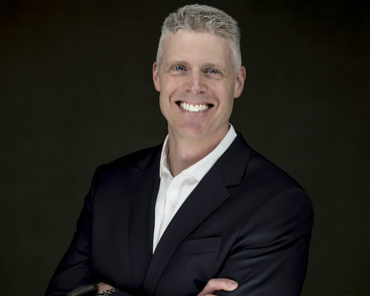 Headshot in MA of man in dark suit jacket and white shirt, smiling warmly against dark background by Rob Faber Photography.