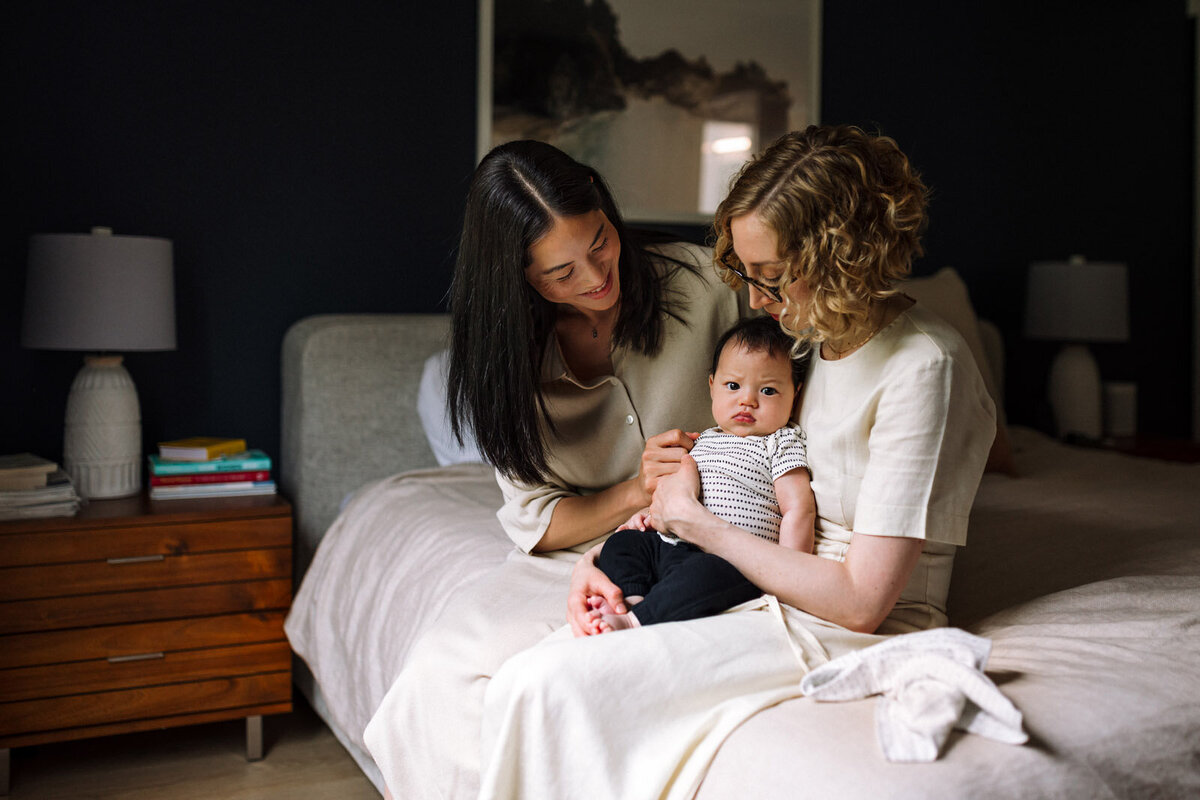 two moms in off-white clothing sitting on neutral bed holding and looking and baby who is looking at camera