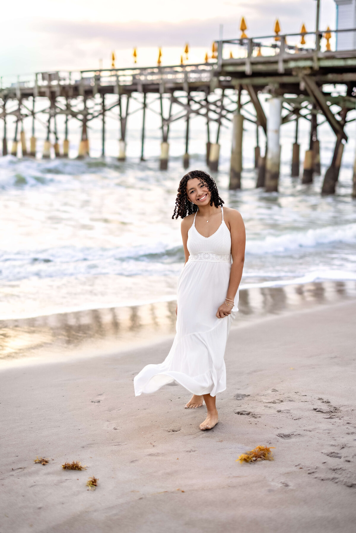Senior girl walking along the sand with Cocoa Beach Pier behind her, waves crashing at her feet and sunlight glowring.