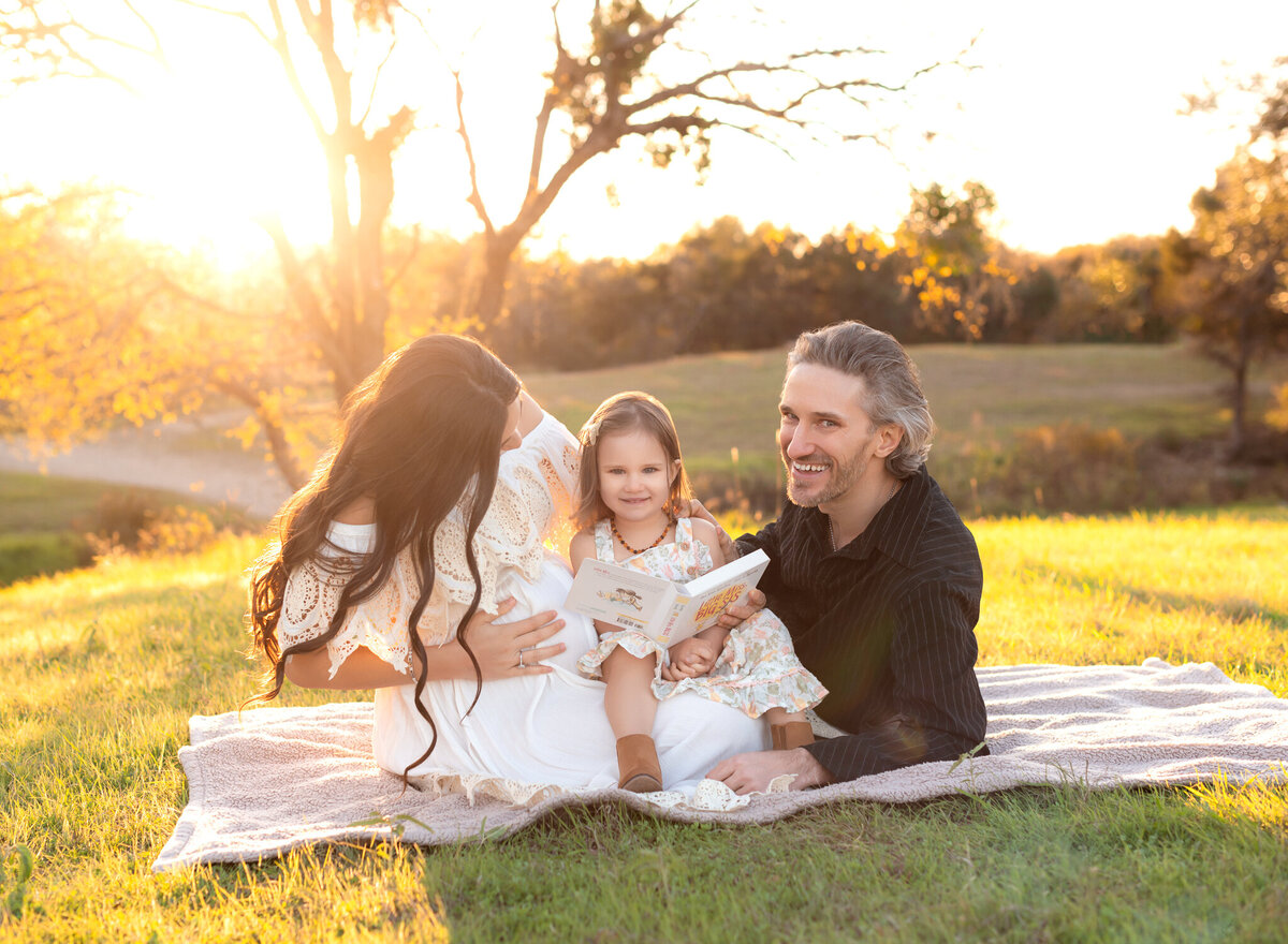 Warm and joyful family maternity session in Leander, Texas, featuring a pregnant mother reading with her daughter and husband during golden hour.