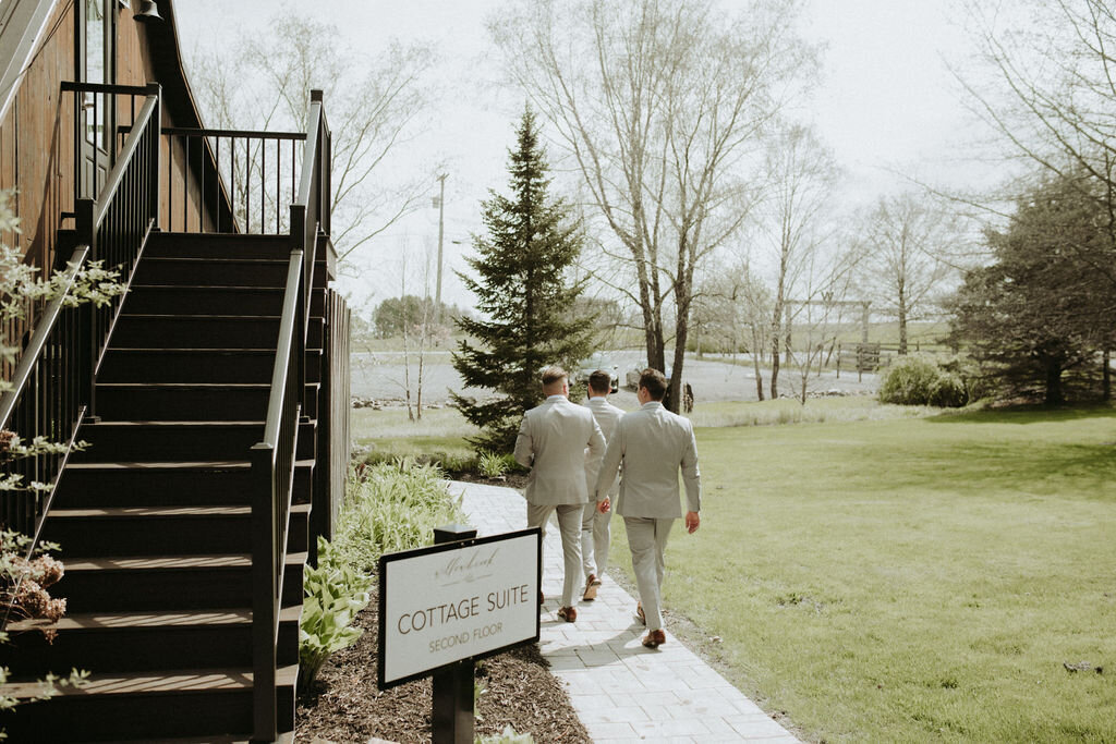 three groomsmen in tan suits walking the stone pathway around the preparation cottage on the grounds of Willowbrook wedding venue in Volant, PA.