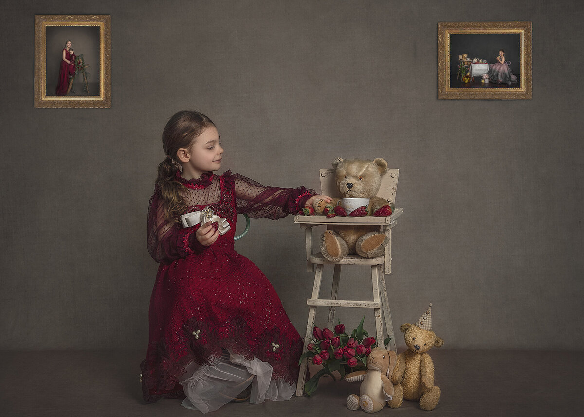 A young girl dressed in red sits beside a teddy bear in a vintage high chair and is touching it's arm gently.
