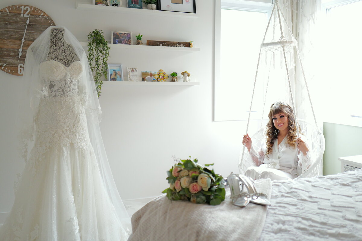 A bride sits gracefully in a swing, with her wedding dress elegantly draped beside her. This image captures a whimsical and charming moment, highlighting the bride’s relaxed elegance and the intricate details of her gown in a beautiful, serene setting.