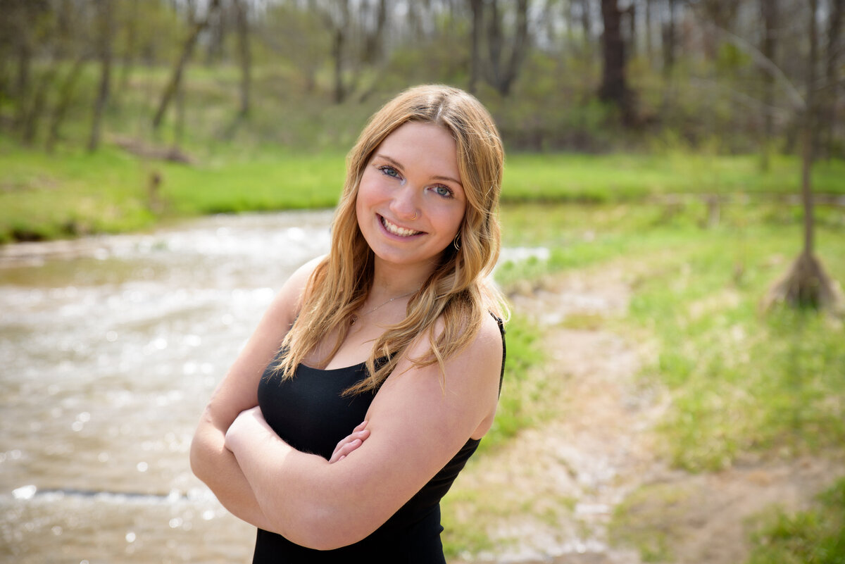 Beautiful high school senior girl with nicely curled hair standing by creek with a black dress on at Fonferek Glen County Park in Green Bay, Wisconsin