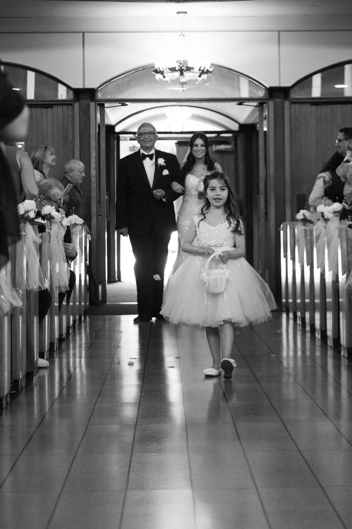 This black and white image features a flower girl walking down the aisle, adding a touch of charm and tradition to the wedding ceremony. The monochrome effect emphasizes the innocence and joy of the moment, focusing on the flower girl as she gracefully makes her way towards the altar.