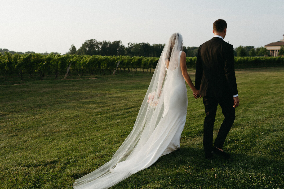 Bride and groom holding hands, walking on grass at Two Sisters Vineyard. The bride wears a white dress with a long veil, and the groom is in a black suit. Captured beautifully by Jennifer van Son, a wedding photographer in Toronto, with trees and a small building visible in the background.