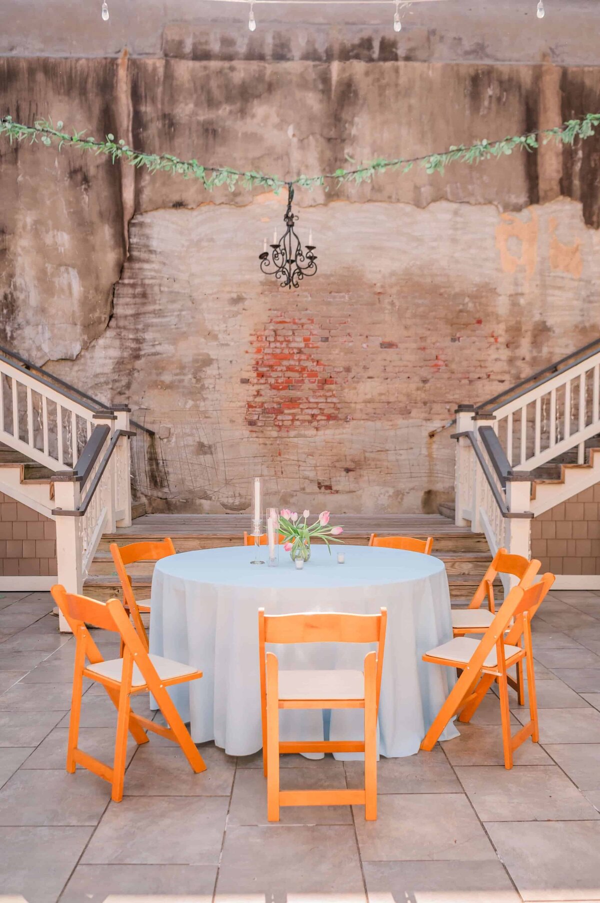 A round table with a light blue tablecloth, a floral centerpiece, and six wooden chairs is set outdoors on a stone courtyard, with greenery and a white railing in the background.