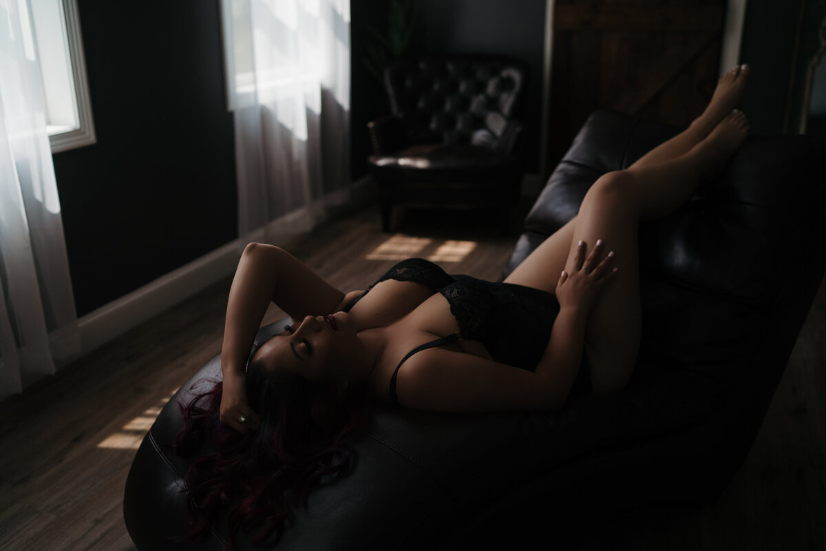 A woman in black lace lingerie lays on a black leather chaise lounge in a studio by windows