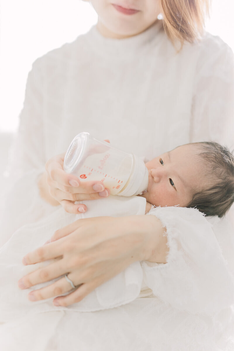 ミルクを飲んでいる新生児の赤ちゃんが、ママの腕に抱かれているナチュラル・ニューボーンフォト。おくるみもママのワンピースも白で統一されたオシャレなスタイリングでYukika Ishikawa Photographyが撮影。