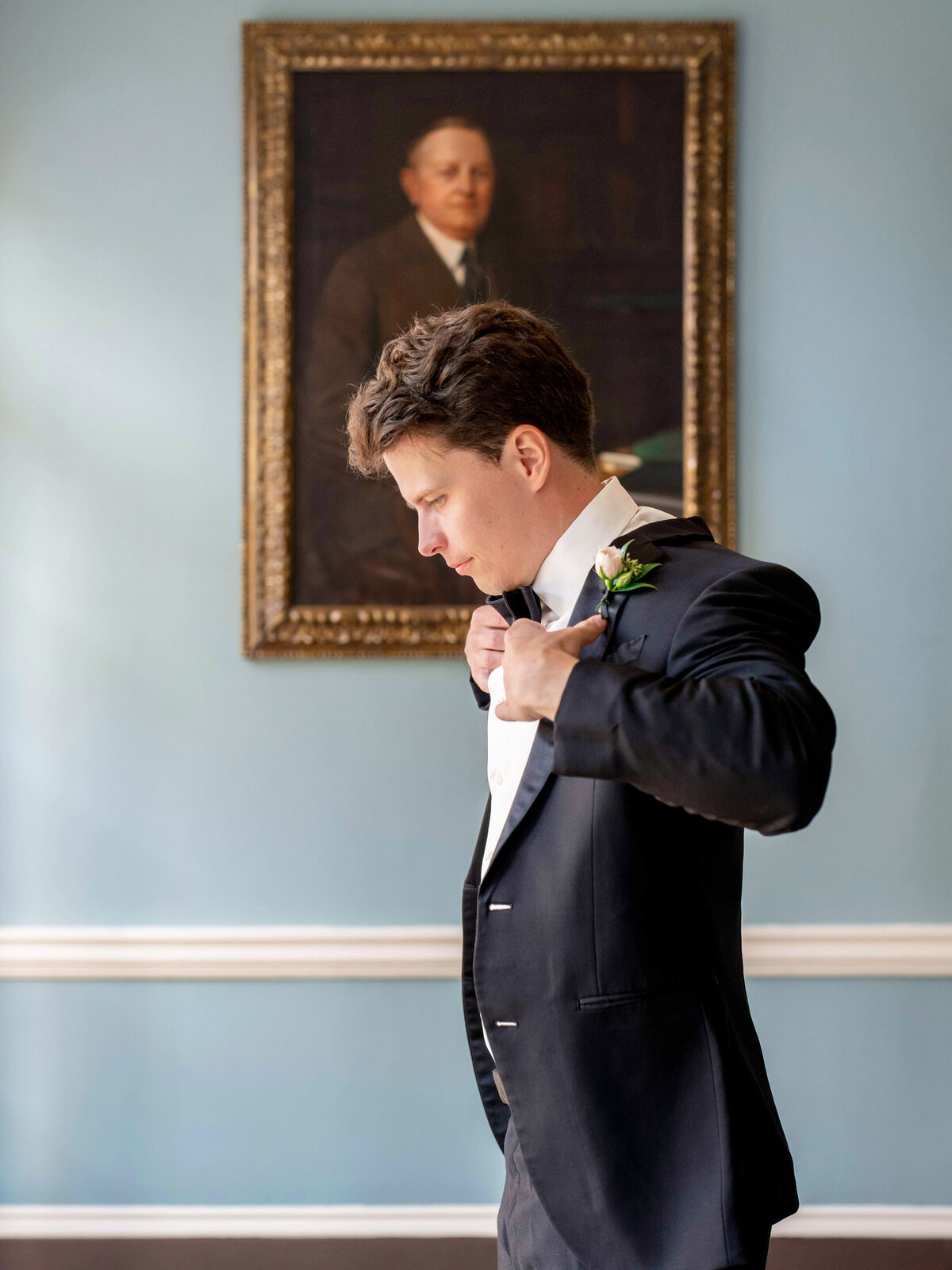 A man in a suit adjusts his bow tie in front of a light blue wall with a large portrait of an older gentleman in a suit hanging in the background. The room has a classic and elegant feel.