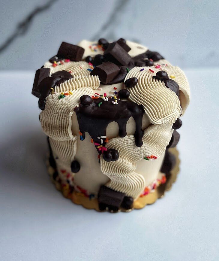 Cake has tan frosting, a chocolate drip, squiggles of frosting, rainbow sprinkles, and chocolate candies and chocolate covered espresso beans decorating it all over.